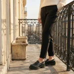 Photo lifestyle d'une femme debout sur un balcon parisien ensoleillé, portant les mocassins noirs à grosse semelle crantée et motif floral gaufré. Elle porte également un pantalon de tailleur noir et un pull en maille crème. La rambarde en fer forgé et l'architecture haussmannienne sont visibles en arrière-plan.