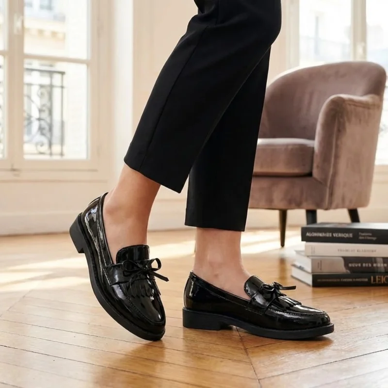 Une vue en gros plan des pieds d'une personne portant des mocassins en cuir verni noir avec des nœuds et des franges. La personne porte un pantalon noir court et se tient debout sur un parquet en bois clair dans une pièce lumineuse et élégante. En arrière-plan, on distingue un fauteuil en velours et une pile de livres près d'une grande fenêtre.