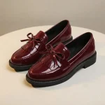 Une paire de mocassins en cuir verni rouge bordeaux brillant pour femme, disposés sur une surface ronde de couleur crème. Comme le modèle noir, ces chaussures possèdent un bout rond, un petit talon, un nœud décoratif et des franges sur le dessus, avec une finition brillante qui capte la lumière.
