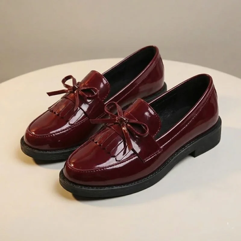 Une paire de mocassins en cuir verni rouge bordeaux brillant pour femme, disposés sur une surface ronde de couleur crème. Comme le modèle noir, ces chaussures possèdent un bout rond, un petit talon, un nœud décoratif et des franges sur le dessus, avec une finition brillante qui capte la lumière.