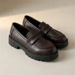 Une paire de mocassins chunky en similicuir marron foncé avec une épaisse semelle crantée noire, posée sur un fond de studio beige uni.