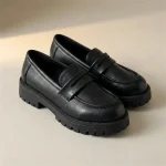 Une paire de mocassins chunky en similicuir noir avec une épaisse semelle crantée noire, posée sur un fond de studio beige uni.