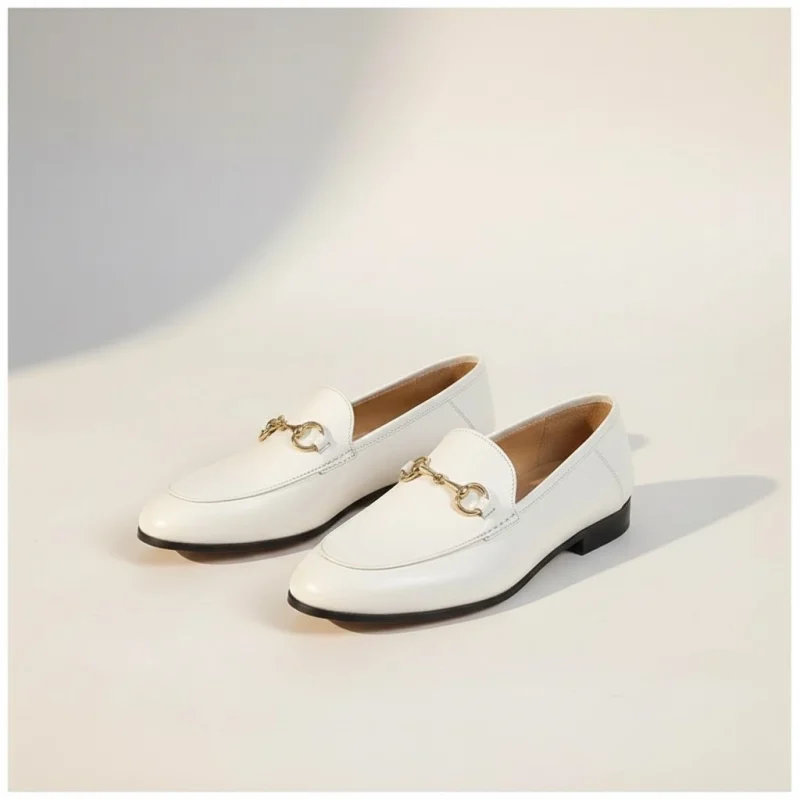 Paire de mocassins blancs en cuir avec un mors doré décoratif, vue de trois quarts sur un fond studio beige chaud.