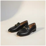 Mocassin Femme en Cuir – Style Britannique noir Paire de mocassins noirs en cuir avec un mors doré décoratif, vue de trois quarts sur un fond studio beige chaud.