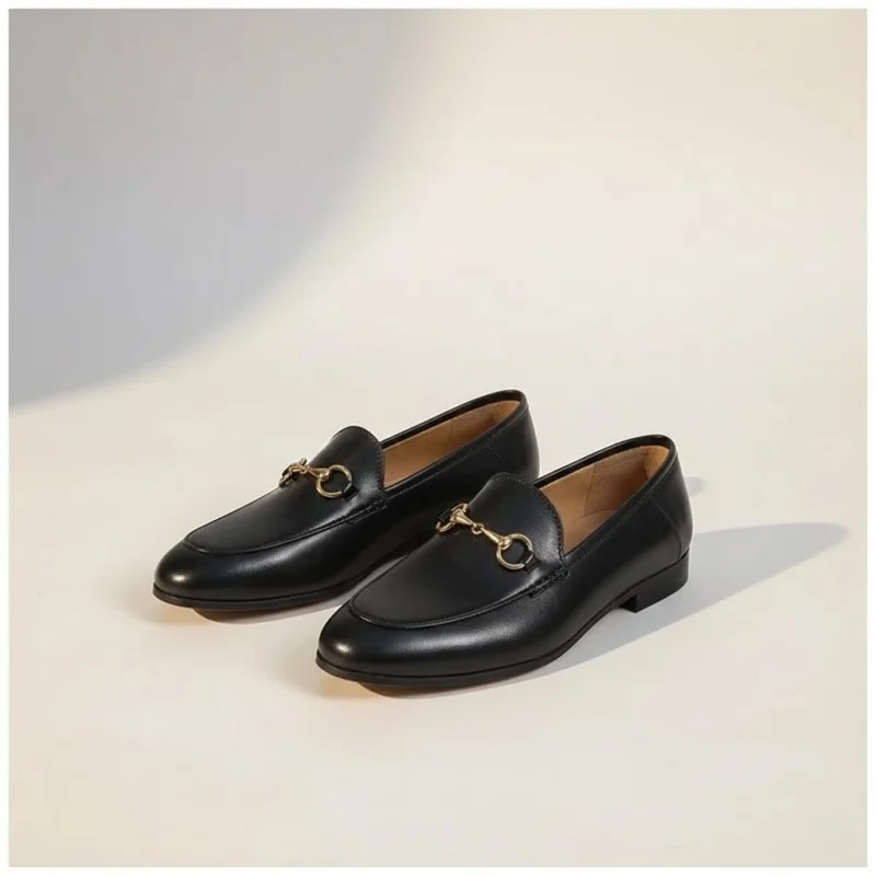 Mocassin Femme en Cuir – Style Britannique noir Paire de mocassins noirs en cuir avec un mors doré décoratif, vue de trois quarts sur un fond studio beige chaud.