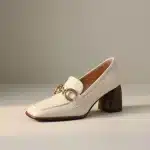 Photo studio d'un mocassin en cuir verni beige (écru/ivoire) avec un talon bloc texturé en bois foncé et une boucle dorée de style mors (horsebit) sur le devant, sur un fond uni.