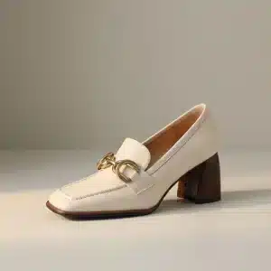 Photo studio d'un mocassin en cuir verni beige (écru/ivoire) avec un talon bloc texturé en bois foncé et une boucle dorée de style mors (horsebit) sur le devant, sur un fond uni.