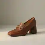Photo studio d'un mocassin en cuir marron chaud (cognac/caramélisé) avec un talon bloc texturé en bois assorti et une boucle dorée de style mors, sur un fond uni