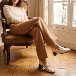 Photo lifestyle d'une femme assise dans un fauteuil en velours ancien dans un appartement parisien. Elle porte un pull blanc, un pantalon camel large et les mocassins beiges à talon bloc et mors doré. Un parquet en chevrons et une fenêtre à la française sont visibles.