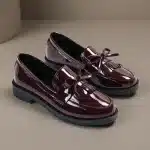 Une paire de mocassins vernis bordeaux profond pour femme avec franges kiltie et nœuds décoratifs, sur un fond de studio gris-beige minimaliste. C'est un plan de référence de produit propre.