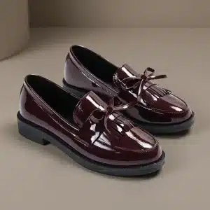 Une paire de mocassins vernis bordeaux profond pour femme avec franges kiltie et nœuds décoratifs, sur un fond de studio gris-beige minimaliste. C'est un plan de référence de produit propre.