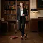 Une paire de mocassins vernis bordeaux profond pour femme avec franges kiltie et nœuds décoratifs, porté par un mannequin en tailleur dans une bibliothèque