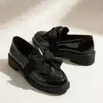 Une paire de mocassins pour femme en cuir verni noir brillant, posée sur une surface de studio claire. Chaque chaussure est ornée d'un grand nœud décoratif en tissu noir mat sur le devant. Elles ont des semelles épaisses et crantées en caoutchouc noir. L'éclairage doux de studio crée des reflets nets sur le cuir verni et des ombres diffuses en arrière-plan.