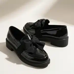 Une paire de mocassins pour femme en cuir verni noir brillant, posée sur une surface de studio claire. Chaque chaussure est ornée d'un grand nœud décoratif en tissu noir mat sur le devant. Elles ont des semelles épaisses et crantées en caoutchouc noir. L'éclairage doux de studio crée des reflets nets sur le cuir verni et des ombres diffuses en arrière-plan.