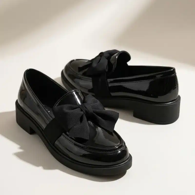 Une paire de mocassins pour femme en cuir verni noir brillant, posée sur une surface de studio claire. Chaque chaussure est ornée d'un grand nœud décoratif en tissu noir mat sur le devant. Elles ont des semelles épaisses et crantées en caoutchouc noir. L'éclairage doux de studio crée des reflets nets sur le cuir verni et des ombres diffuses en arrière-plan.