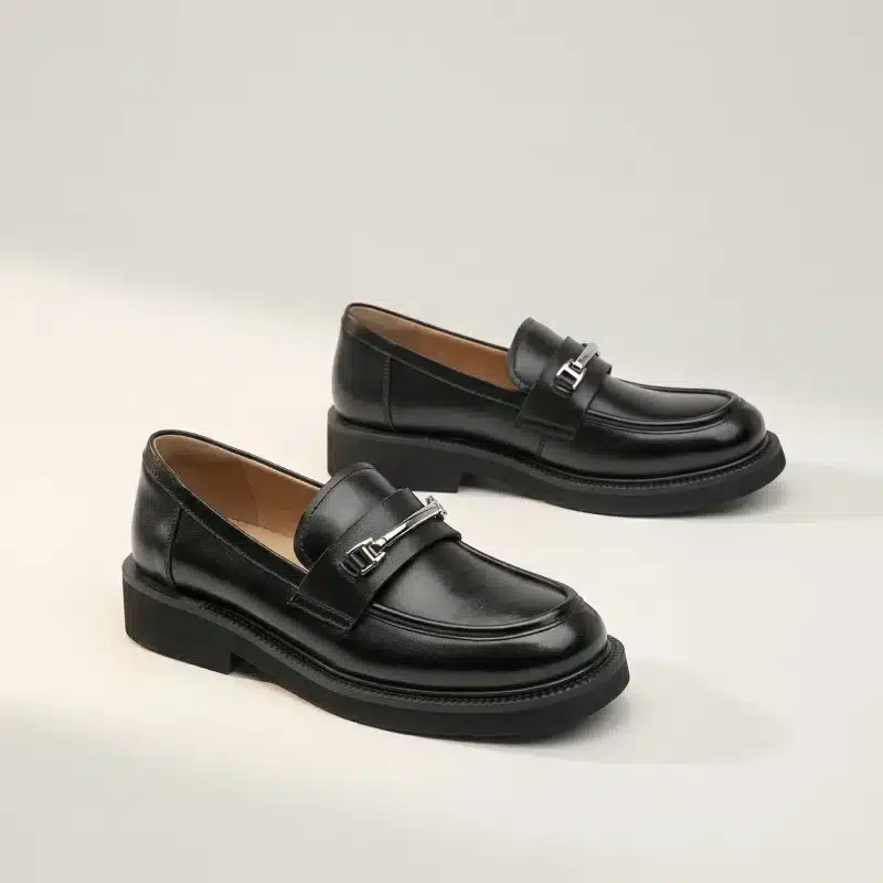 Photo de produit d'une paire de mocassins en cuir noir à semelle crantée et boucle argentée en forme de H, présentés sur un fond de studio warm off-white.