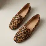 Photographie de produit de haute qualité, vue de dessus, d'une paire de mocassins à motif léopard texturé et boucles mors de cheval dorées sur un fond de studio gris-blanc chaud uni, avec un éclairage doux et directionnel du coin supérieur gauche et une profondeur de champ faible.