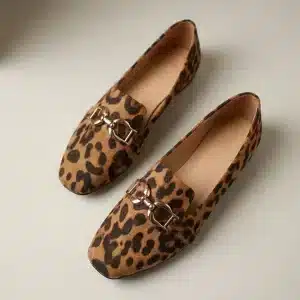 Photographie de produit de haute qualité, vue de dessus, d'une paire de mocassins à motif léopard texturé et boucles mors de cheval dorées sur un fond de studio gris-blanc chaud uni, avec un éclairage doux et directionnel du coin supérieur gauche et une profondeur de champ faible.