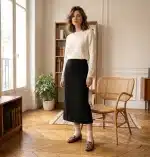 Photographie de mode en pied d'une mannequin européenne debout dans un appartement parisien élégant, portant un pull ivoire, une jupe noire, des chaussettes crème et les mocassins léopard avec boucles dorées.