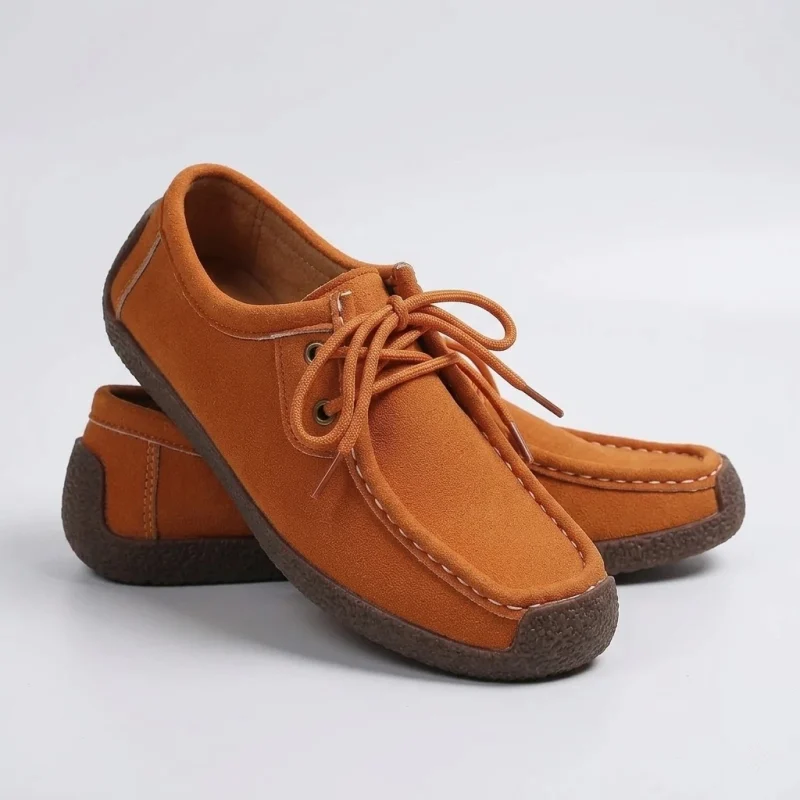 Paire de mocassins plats en daim orange vif avec semelle marron texturée, lacet assorti et surpiqûres blanches contrastées. Présentation de produit de studio sur fond gris clair et blanc.