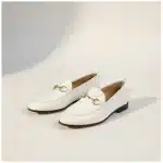 Mocassin Femme en Cuir – Style Britannique blanc Paire de mocassins blancs en cuir avec un mors doré décoratif, vue de trois quarts sur un fond studio beige chaud.
