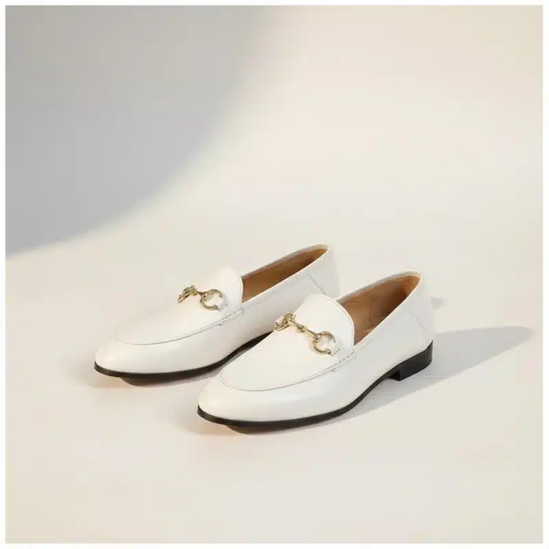 Paire de mocassins blancs en cuir avec un mors doré décoratif, vue de trois quarts sur un fond studio beige chaud.