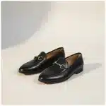 Mocassin Femme en Cuir – Style Britannique noir Paire de mocassins noirs en cuir avec un mors doré décoratif, vue de trois quarts sur un fond studio beige chaud.