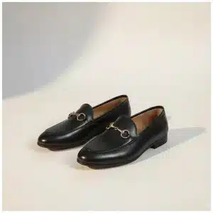 Paire de mocassins noirs en cuir avec un mors doré décoratif, vue de trois quarts sur un fond studio beige chaud.