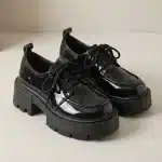 Photo de produit d'une paire de chaussures Derby noires en cuir verni brillant, sur un fond blanc cassé. Les chaussures présentent une semelle plateforme épaisse et crantée, des lacets noirs et une languette à l'arrière.