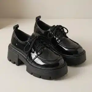 Photo de produit d'une paire de chaussures Derby noires en cuir verni brillant, sur un fond blanc cassé. Les chaussures présentent une semelle plateforme épaisse et crantée, des lacets noirs et une languette à l'arrière.