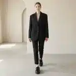 Une femme dans un ensemble tailleur noir, composé d'un blazer oversized et d'un pantalon slim, marche vers l'appareil photo dans un studio lumineux et minimaliste. Elle porte des chaussures Derby noires en cuir verni avec une semelle plateforme épaisse et crantée.
